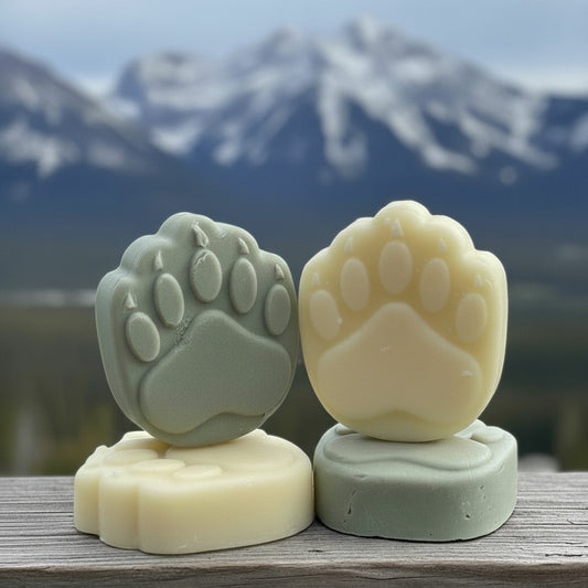 Two soap bars shaped like paw prints on a wooden surface with a mountainous landscape in the background.