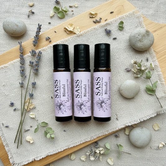 Three bottles of SASS Blissful roll-on essential oil on a wooden cutting board with lavender on the side and a couple rocks placed by 
